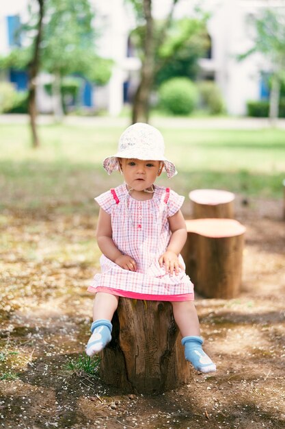 Petite fille au chapeau est assise sur une souche d'arbre dans la cour