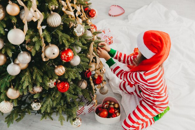 Petite fille au chapeau de père Noël décore l'arbre avec des jouets. Un bébé mignon se prépare à fêter Noël.