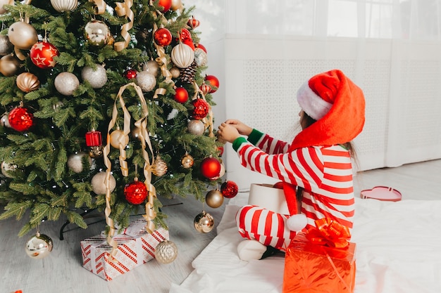 Petite fille au chapeau de père Noël décore l'arbre avec des jouets. Un bébé mignon se prépare à fêter Noël.