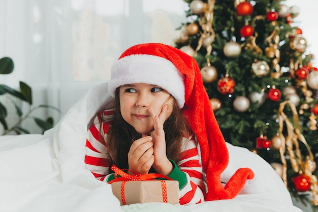 Petite fille au chapeau rouge du Nouvel An avec un cadeau de Noël. Elle est allongée sur le lit, serrant dans ses bras une boîte sur fond d'arbre de Noël.