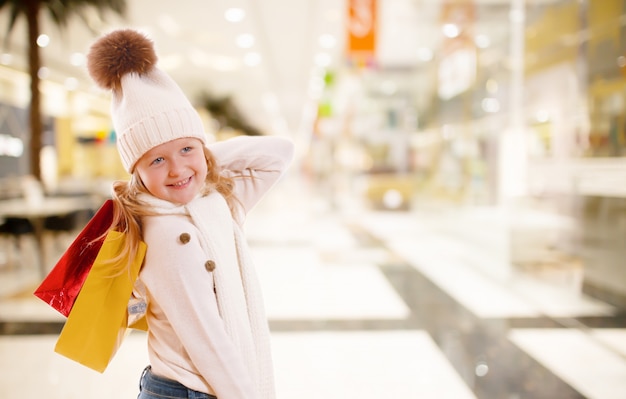 Une petite fille au chapeau tricoté tient des sacs en papier multicolores dans un centre commercial.