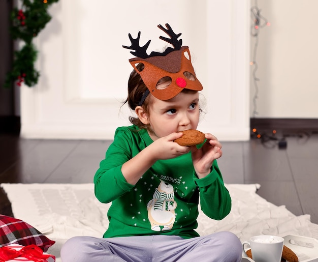 Petite fille au masque de cerf drôle et pyjama de Noël assis sur un plaid mangeant des biscuits et buvant du cacao