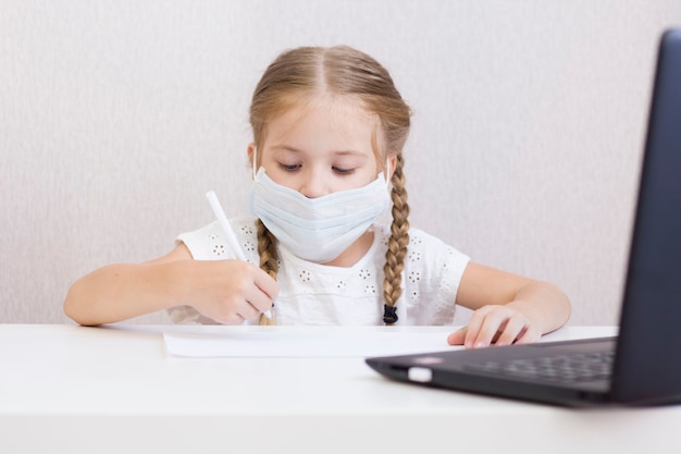 UNE PETITE FILLE AU MASQUE EST ASSIS À UNE TABLE ET TIRE, ÉCRIRE. ÉTUDIANT. L'ÉCOLE. LE PREMIER SEPTEMBRE. QUARANTAINE À DOMICILE