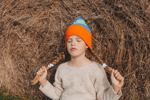 Une petite fille aux beaux cheveux blonds est assise sur un champ près d'un rouleau de foin. portrait d'un enfant dans un bonnet tricoté.