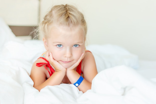 Une petite fille aux cheveux blonds est allongée dans un lit avec des draps blancs.