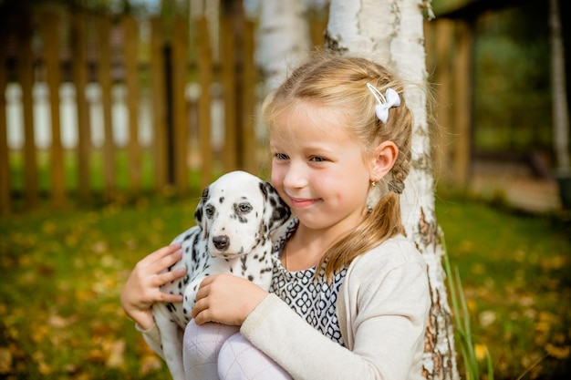 Petite fille aux cheveux blonds joue avec un chiot sur un fond de jardinPetite fille tient un chiot sur ses brasMignonne petite fille étreignant un chiot dalmatien