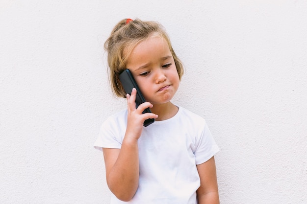 Petite fille aux cheveux blonds portant un t-shirt blanc, parlant au téléphone portable, avec un visage mécontent