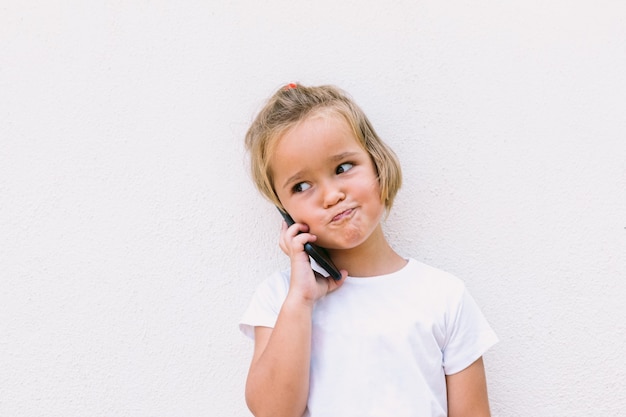 Petite fille aux cheveux blonds portant un t-shirt blanc, parlant au téléphone portable, avec un visage souriant