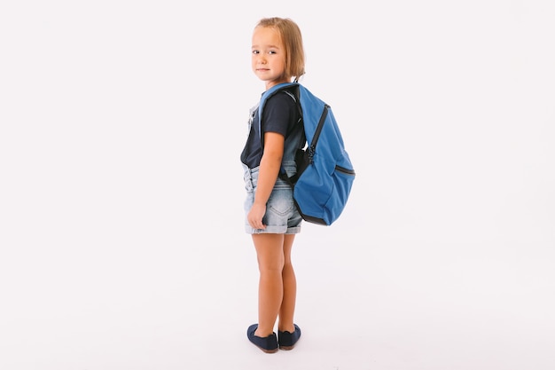 Petite Fille Aux Cheveux Blonds Vêtue D'une Salopette En Jean Et D'un T-shirt Bleu, Avec Un Sac à Dos Prêt Pour La Rentrée, De Son Côté, Sur Fond Blanc