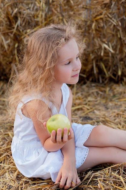 une petite fille aux cheveux bouclés est assise près d'une botte de foin dans un champ avec une pomme verte dans les mains