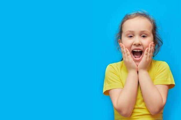 Une petite fille aux cheveux bouclés ouvre grand la bouche surprise sur fond bleu