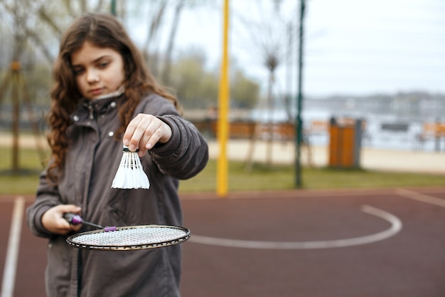 Photo petite fille aux cheveux bruns bouclés tenant un volant et se préparant à un match de badminton dans la cour de sport. espace pour le texte