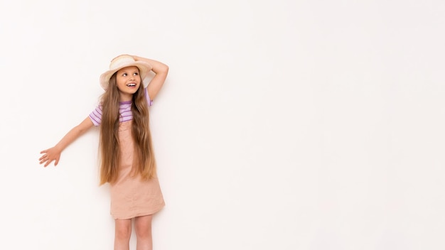 Une petite fille aux cheveux longs dans une robe d'été et un chapeau de paille s'appuie contre un mur blanc et regarde votre publicité en souriantCopier l'espace