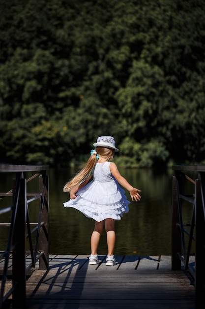 Une petite fille aux cheveux longs vêtue d'une robe blanche se dresse au bord de la rivière. Promenade au bord du lac par une journée ensoleillée d'été