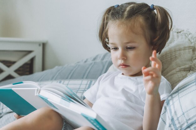 Petite Fille Aux Cheveux Noirs 2-4 Avec Deux Queues De Cheval Est Assise Sur Le Lit Avec Des Oreillers Et Lit Un Livre