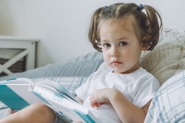 Petite fille aux cheveux noirs 24 avec deux queues de cheval est assise sur le lit avec des oreillers et lit un livre