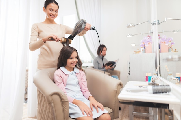 Une petite fille aux cheveux noirs est assise dans le fauteuil du salon de beauté