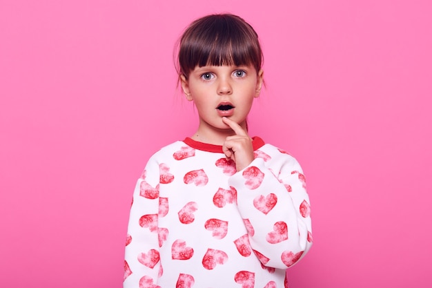 Petite fille aux cheveux noirs avec une frange regarde la caméra avec de grands yeux surpris, tient son doigt sur sa bouche, robes pull, isolé sur un mur rose.