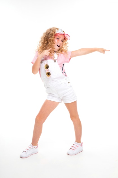 Une petite fille aux cheveux rouges dans une chemise, une combinaison blanche, des baskets blanches, une casquette et des lunettes de soleil rondes danse.