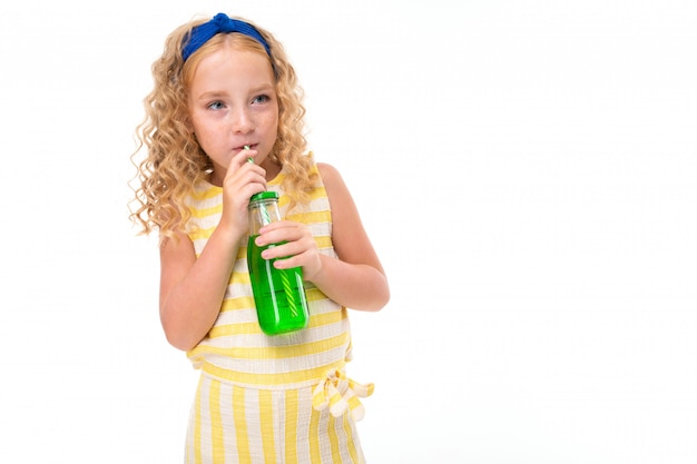 Une petite fille aux cheveux rouges dans un costume d'été à rayures blanches et jaunes, avec un bandage bleu sur la tête, boit du jus d'orange dans une bouteille en verre.