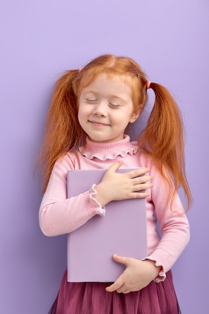 Petite fille aux cheveux rouges isolée. douce petite fille dans un livre de câlins de vêtements décontractés, les yeux fermés. école, concept d'éducation.