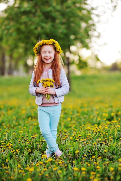 Une petite fille aux cheveux rouges sourit contre un champ de pissenlits et d'herbe verte. Été, enfance, vacances