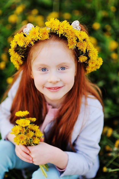 Une petite fille aux cheveux rouges sourit contre un champ de pissenlits et d’herbe verte.