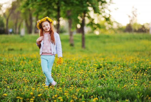 Une petite fille aux cheveux rouges sourit contre un champ de pissenlits et d’herbe verte.