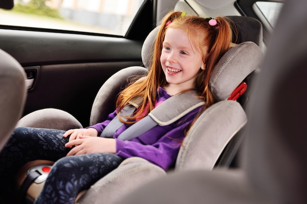 Petite fille aux cheveux roux souriante assise dans un siège auto.