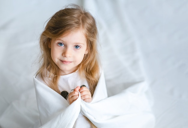 Une petite fille aux longs cheveux blonds était enveloppée dans une couverture blanche sur le lit