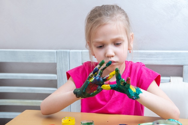 Petite fille aux mains peintes colorées. Une fille concentrée étale de la peinture sur ses mains et regarde le résultat.