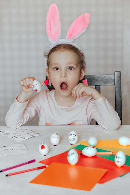 une petite fille aux oreilles de lapin sur la tête décore des œufs blancs avec des autocollants aux émotions différentes