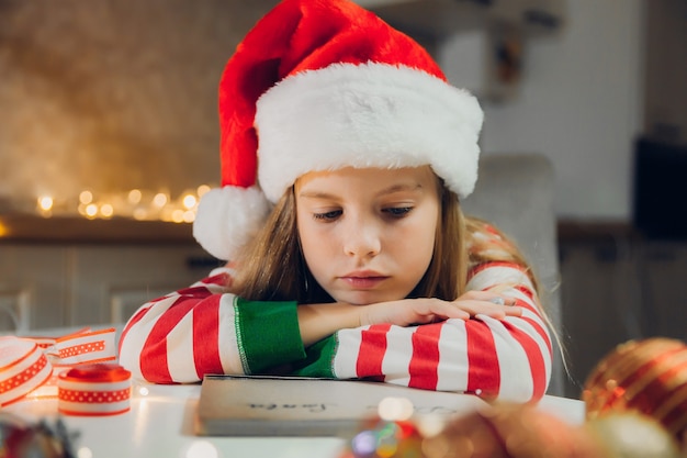 Une petite fille avant Noël écrit une lettre au Père Noël sur la table avec des guirlandes. l'enfant écrit une lettre au père noël.