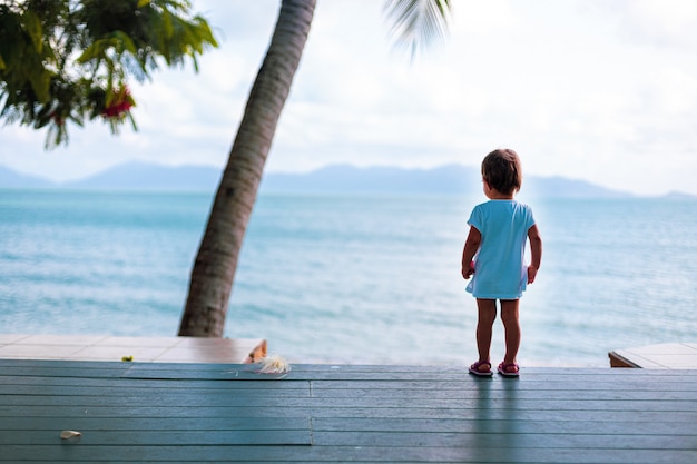 Petite fille en bas âge dans une robe blanche regarde la mer en se tenant debout sur le plancher en bois à l'extérieur