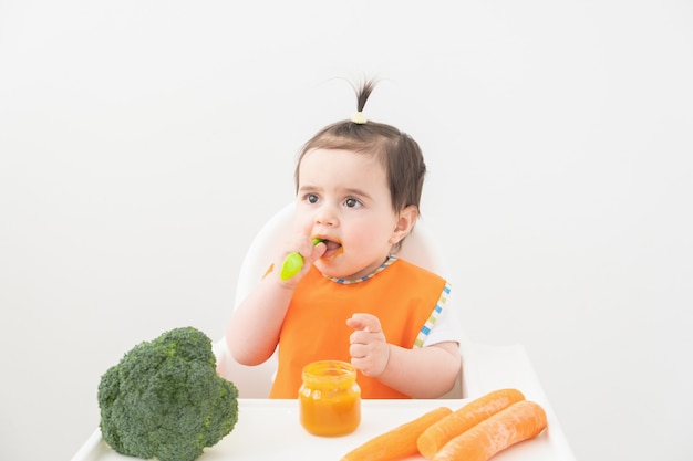 Petite fille en bavoir orange assis dans une chaise d'enfant de manger de la purée de légumes sur fond blanc.