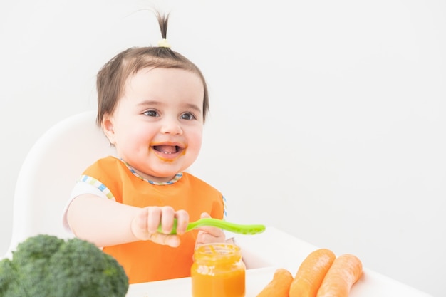 Petite fille en bavoir orange assis dans une chaise d'enfant de manger de la purée de légumes.