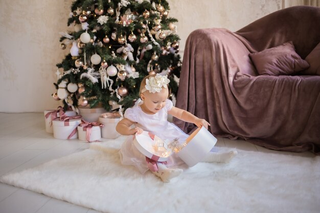 Une petite fille belle dans une robe blanche festive est assise sur une couverture près de l'arbre de Noël et joue avec un cadeau