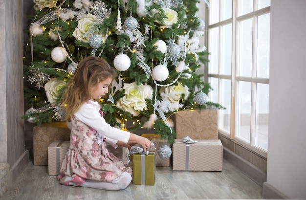 Petite fille en belle robe est assise sous l'arbre de Noël avec boîte-cadeau et arc. Lumière d'une grande fenêtre, pièce sombre, confort de la maison, Noël, magie et joie.