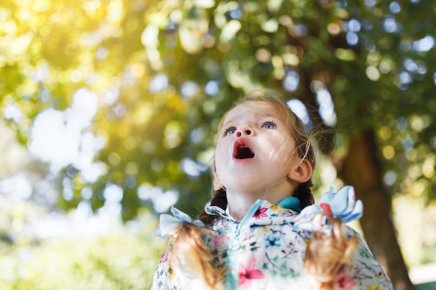 Une petite fille blanche heureuse avec deux nattes dans une veste multicolore surprise que l'automne soit venu et qu'il est temps d'aller à l'école par une chaude journée d'automne