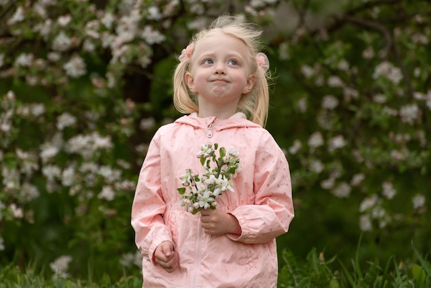 Petite fille blanche joyeuse avec deux queues dans une veste rose dans le jardin de printemps Enfant d'âge préscolaire près d'un arbre en fleurs