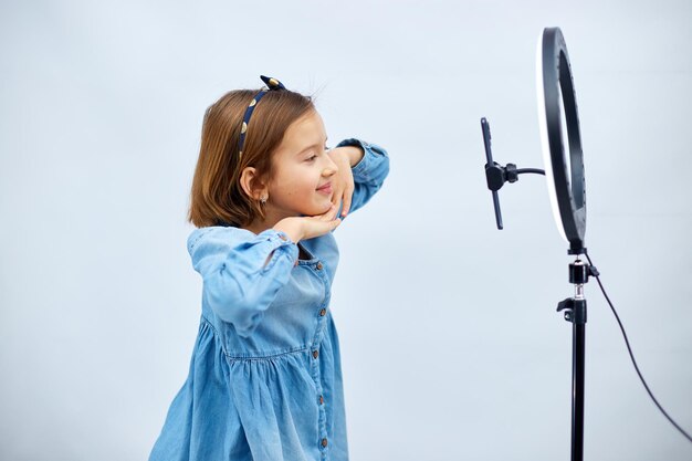 Une petite fille blogueuse souriante en robe denim décontractée utilise une lampe à led selfie et un smartphone