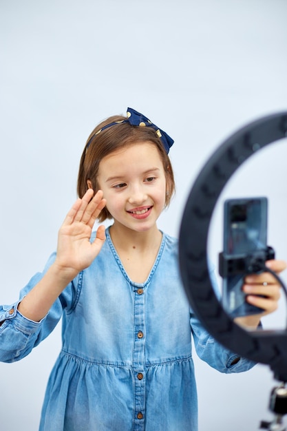 Une petite fille blogueuse souriante en robe denim décontractée utilise une lampe à led selfie et un smartphone