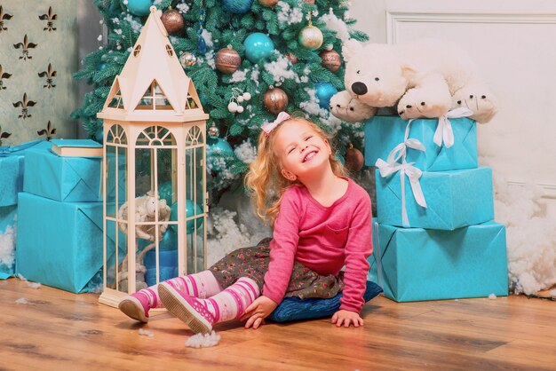 Petite fille blonde assise et souriante presque décorée d'arbre de noël et de cadeaux de nouvel an