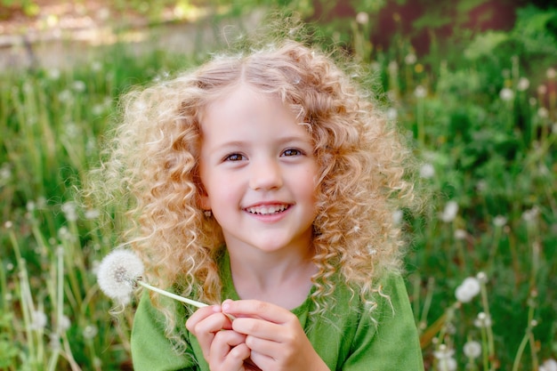 Une petite fille blonde aux cheveux bouclés souffle sur les pissenlits dans une clairière