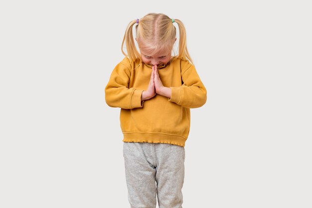 Une petite fille blonde avec une capuche jaune prie sur un fond blanc isolé.