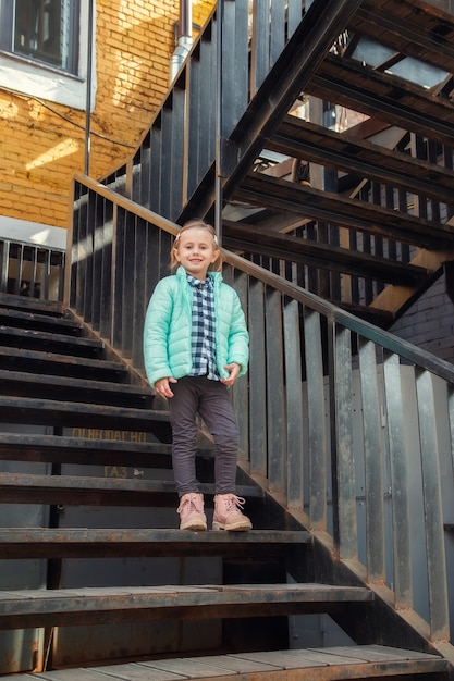 Petite fille blonde caucasienne dans un vêtement de printemps chaud pour les jeunes à la mode se dresse sur l'escalier en fer vintage en regardant la caméra. Jolie belle fille dans des vêtements élégants.