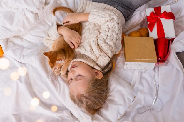 petite fille blonde dans un chandail tricoté est allongée dans son lit avec un chat rouge et des cadeaux pour le Nouvel An,