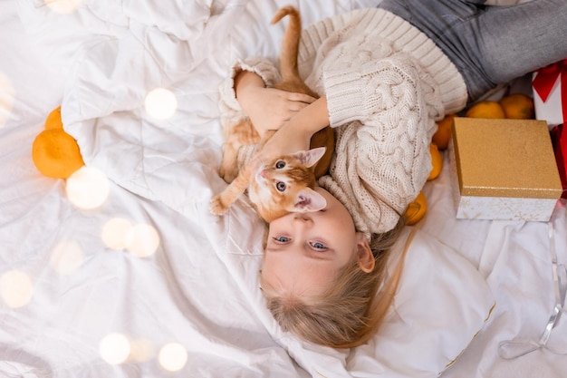 petite fille blonde dans un chandail tricoté est allongée dans son lit avec un chat rouge et des cadeaux pour le Nouvel An,