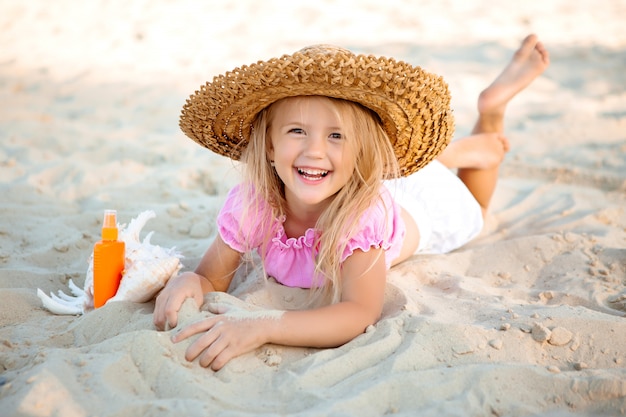 petite fille blonde dans un chapeau de paille est allongé sur une plage de sable à côté d'un coquillage et d'un écran solaire, de l'espace pour le texte