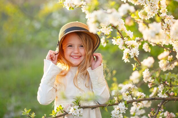 Une petite fille blonde dans un chapeau de paille près d'un arbre en fleurs